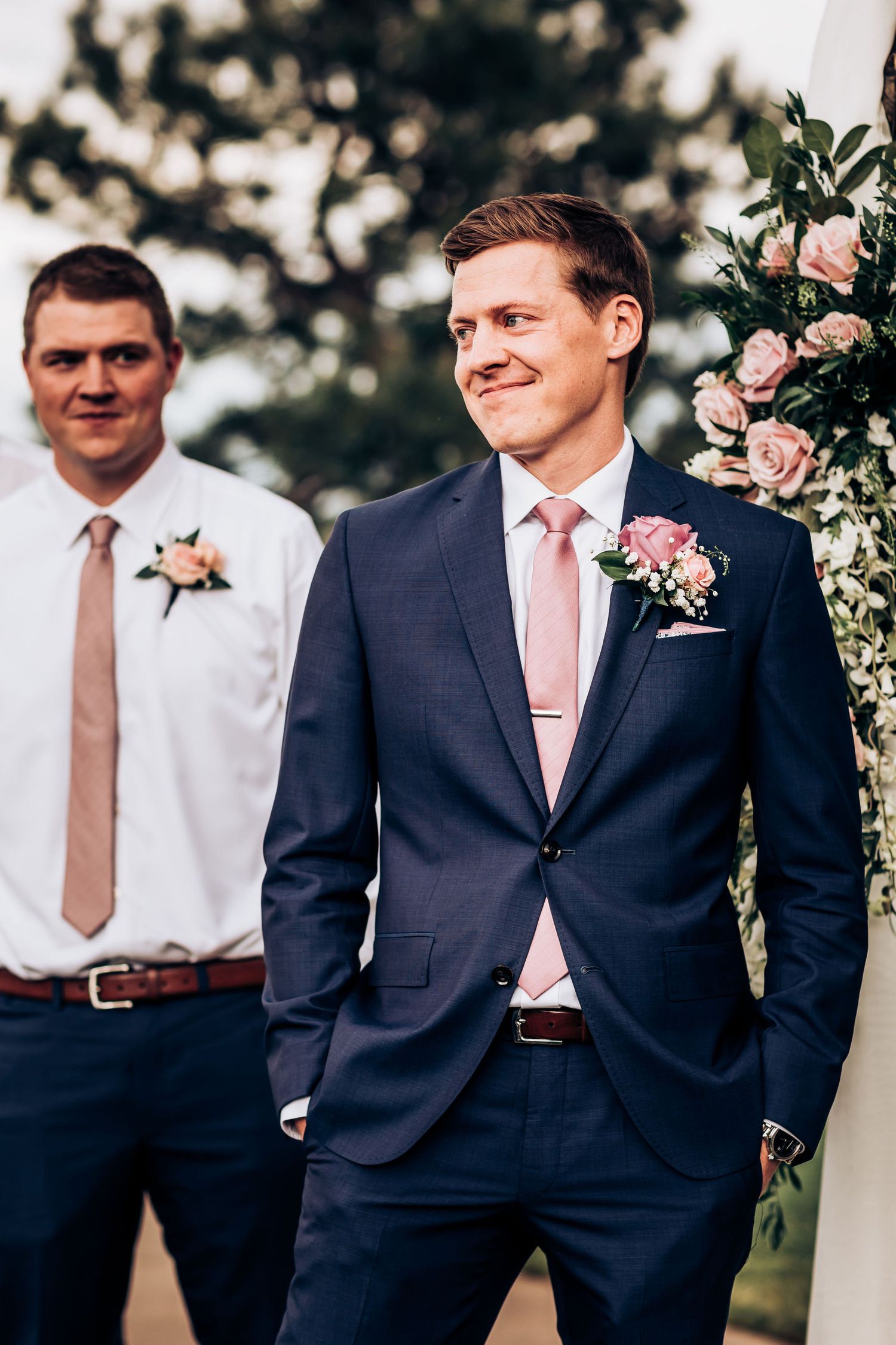 Groomsmen in navy suits with pink accents stand at outdoor wedding venue.