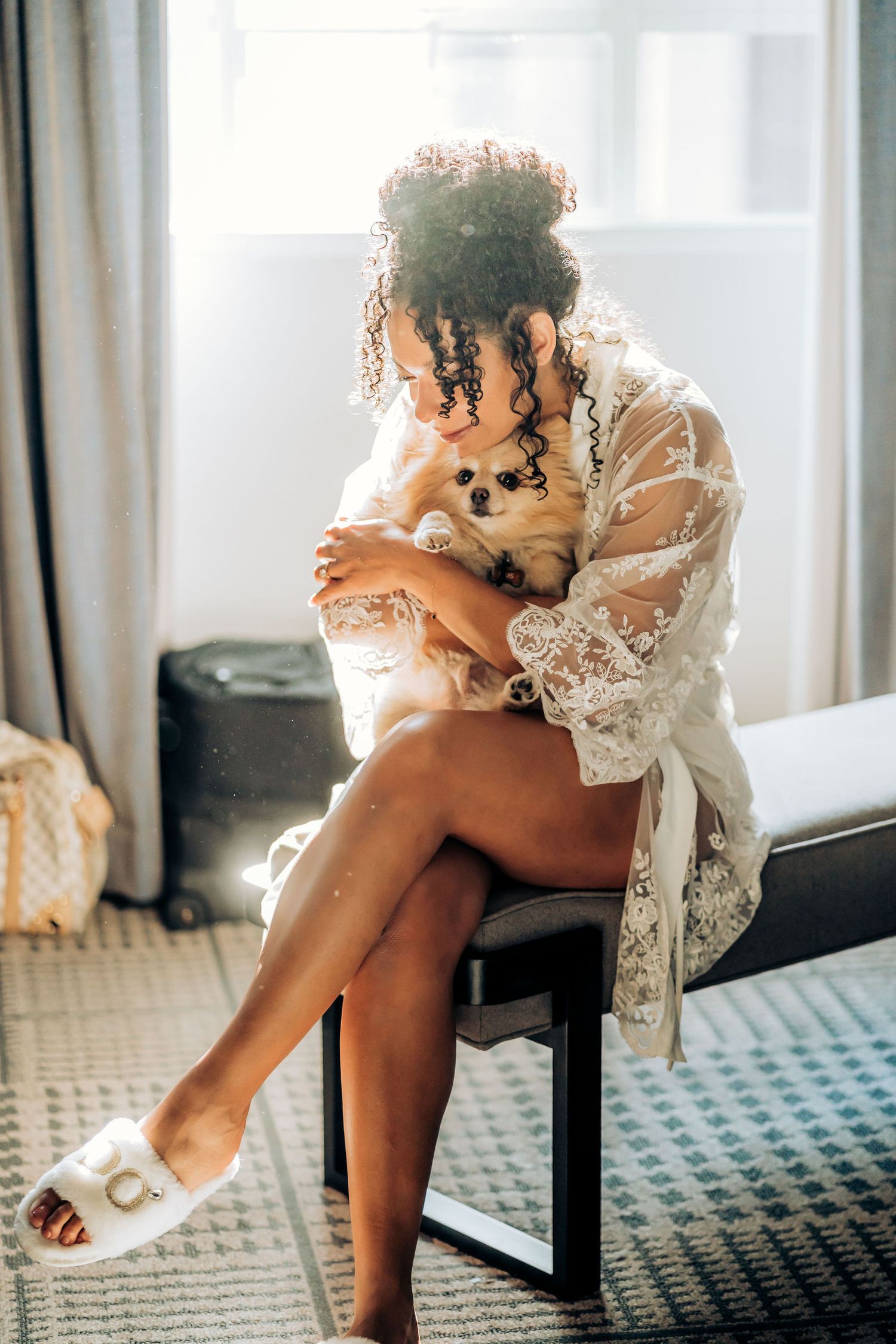 Intimate bridal preparation moment in white lace robe with soft window lighting.