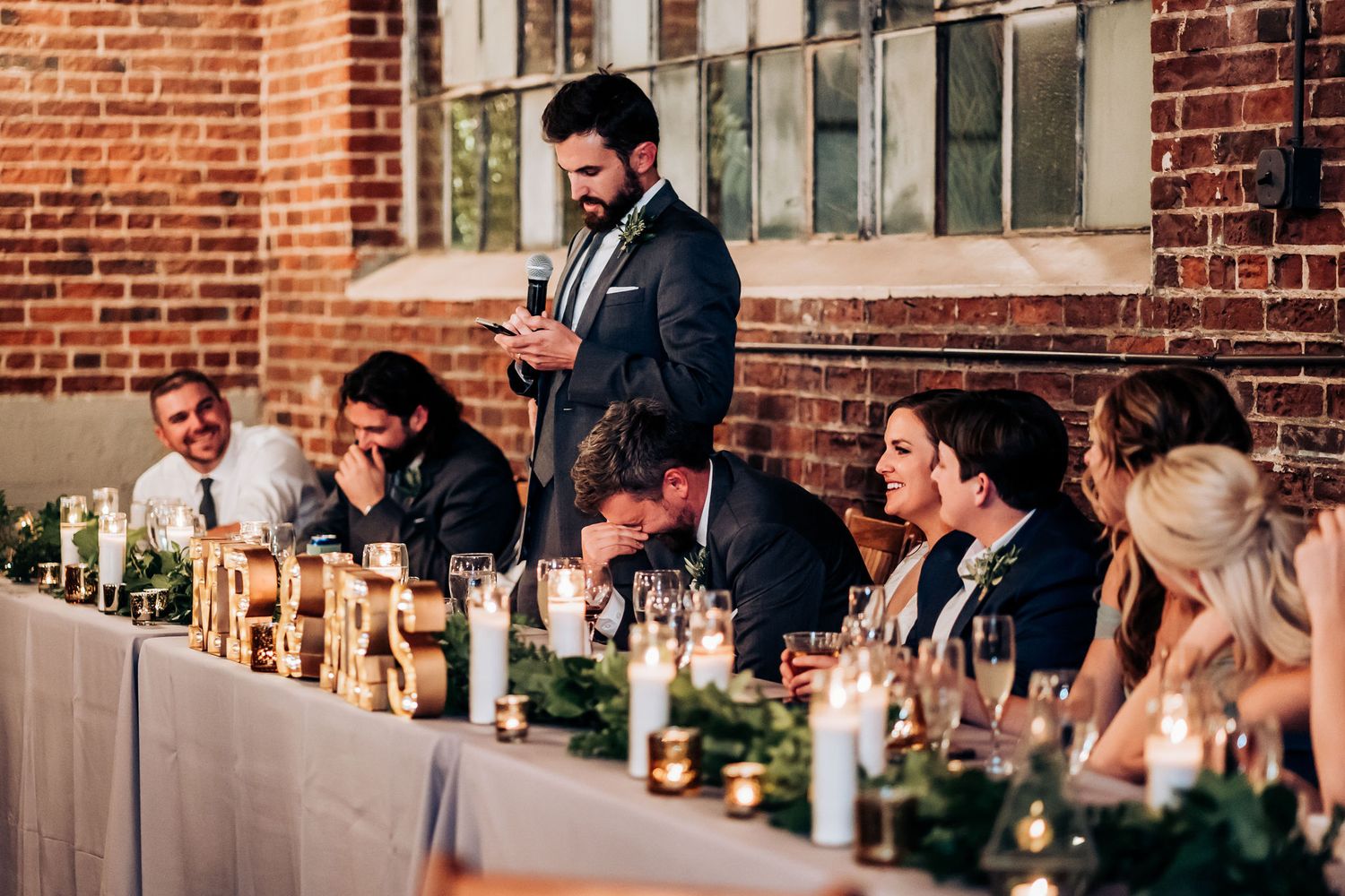 Speaker delivering toast at wedding head table with greenery and candle decorations.