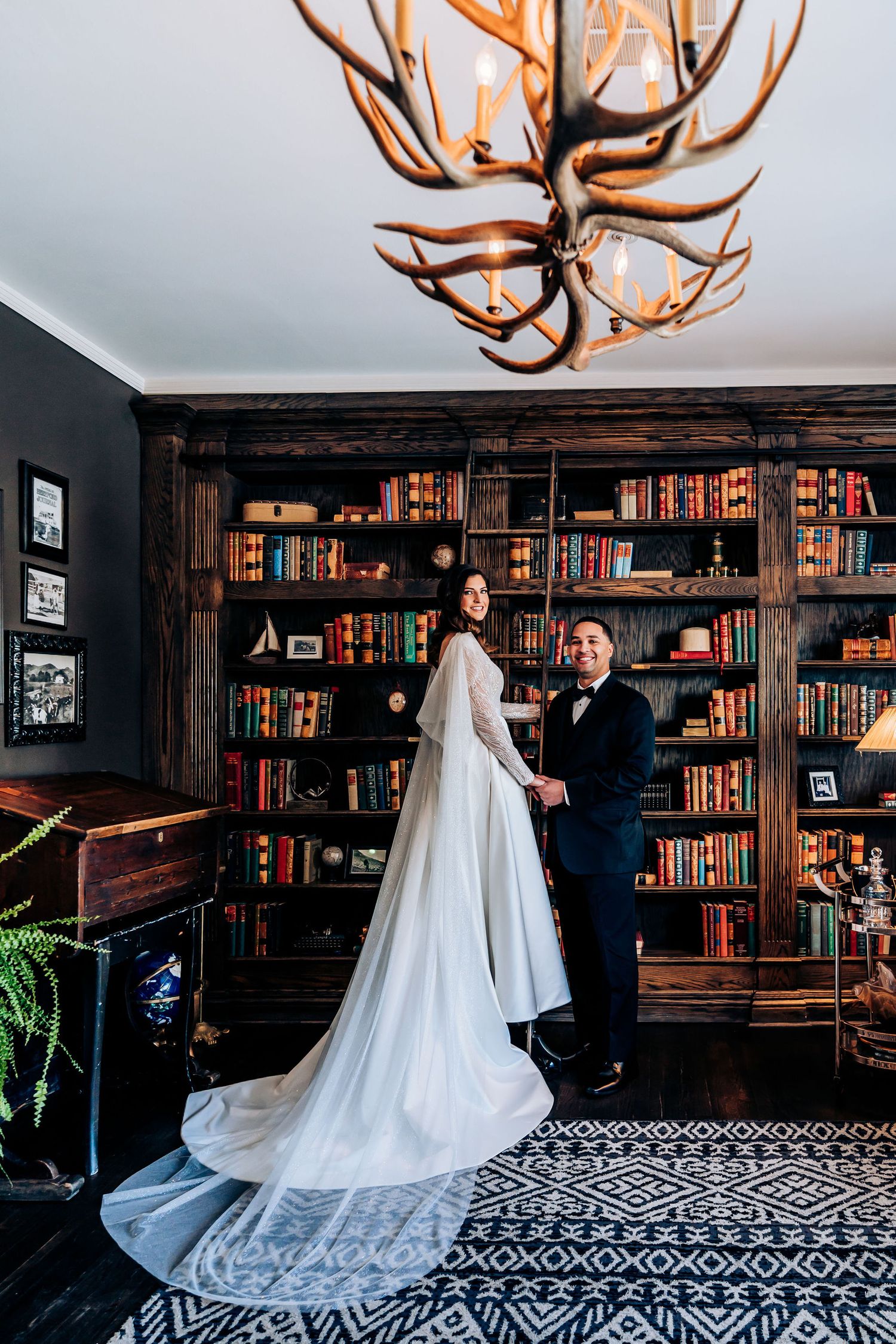 A wedding couple poses in front of a vintage library with antler chandelier and dark bookshelves.