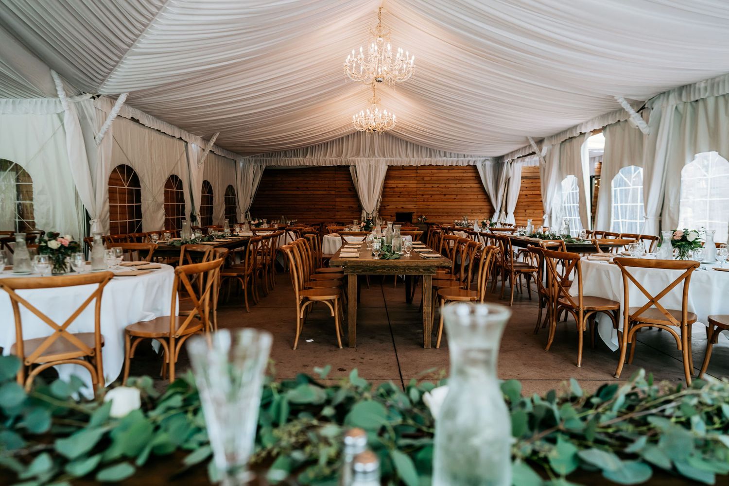 An elegant white tent reception space with wooden cross-back chairs and greenery table decorations.