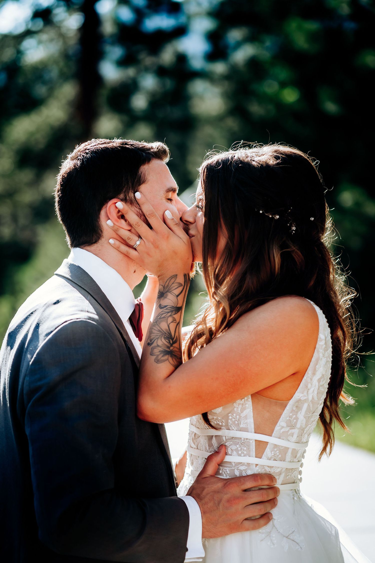 A tender wedding kiss captured in natural sunlight with a forest backdrop.