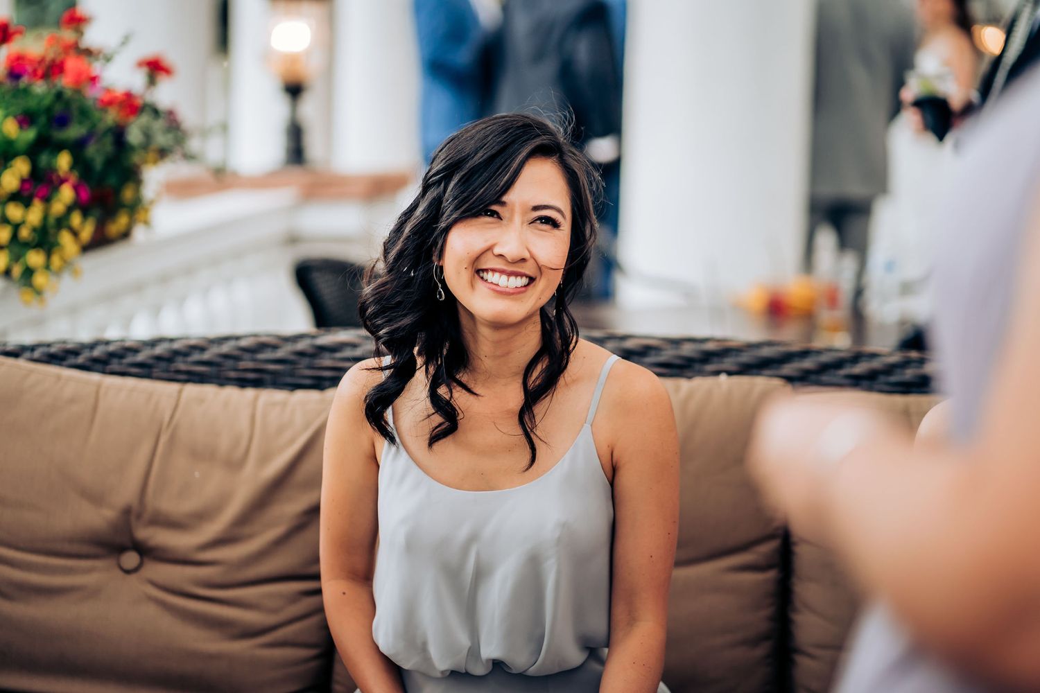 Woman in grey tank top sits smiling on brown couch during sunny outdoor gathering.