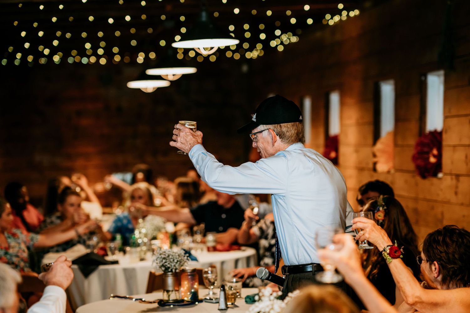 People celebrate at a reception under string lights in a rustic indoor venue.