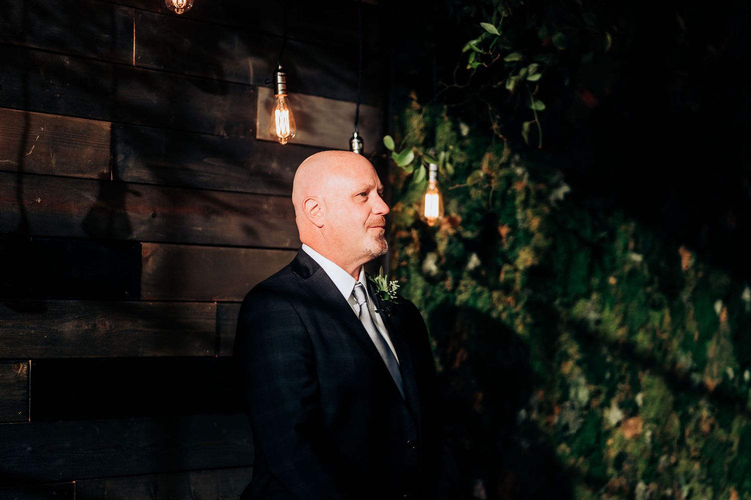 Elegant man in formal attire standing against a dark wooden wall with hanging lights and greenery.
