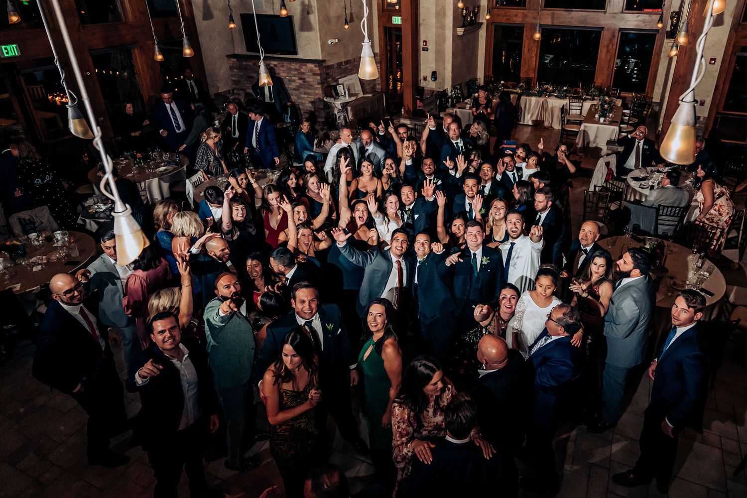A large group of wedding guests gathered on the dance floor during an indoor reception.