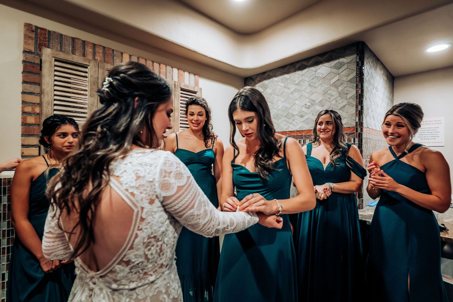 Wedding party in teal dresses gathers around bride wearing lace sleeve gown.