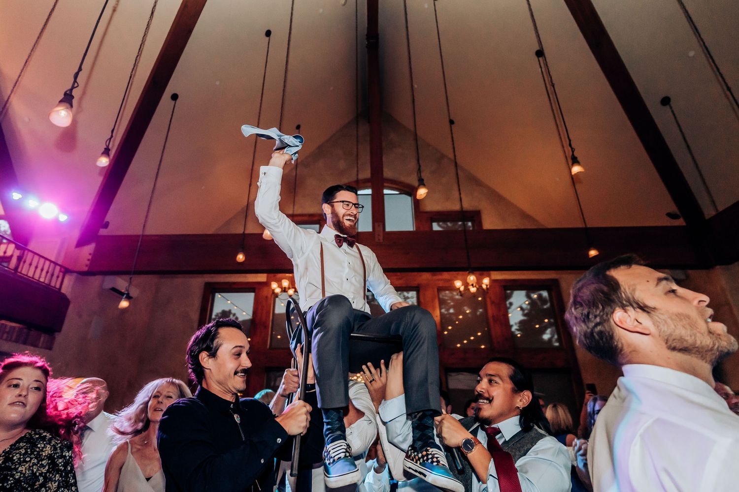 A celebratory moment at a wedding reception shows guests lifting someone high on their shoulders.