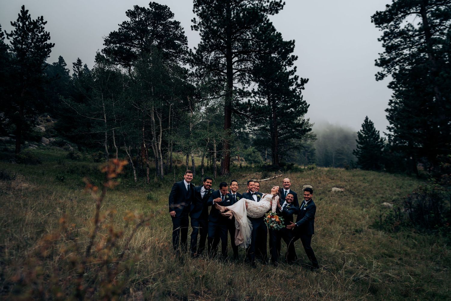 Wedding party carrying couple on hillside with tall evergreen trees in mountain forest setting.