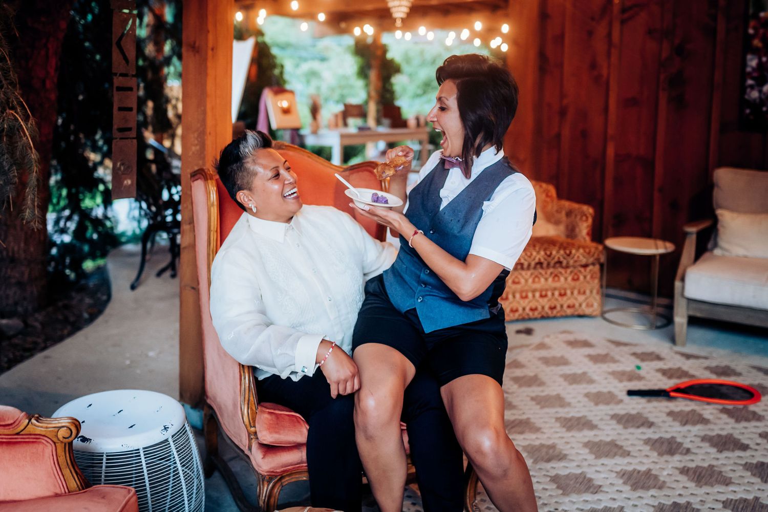 Two people sharing laughter while sitting together in a cozy indoor setting with string lights.
