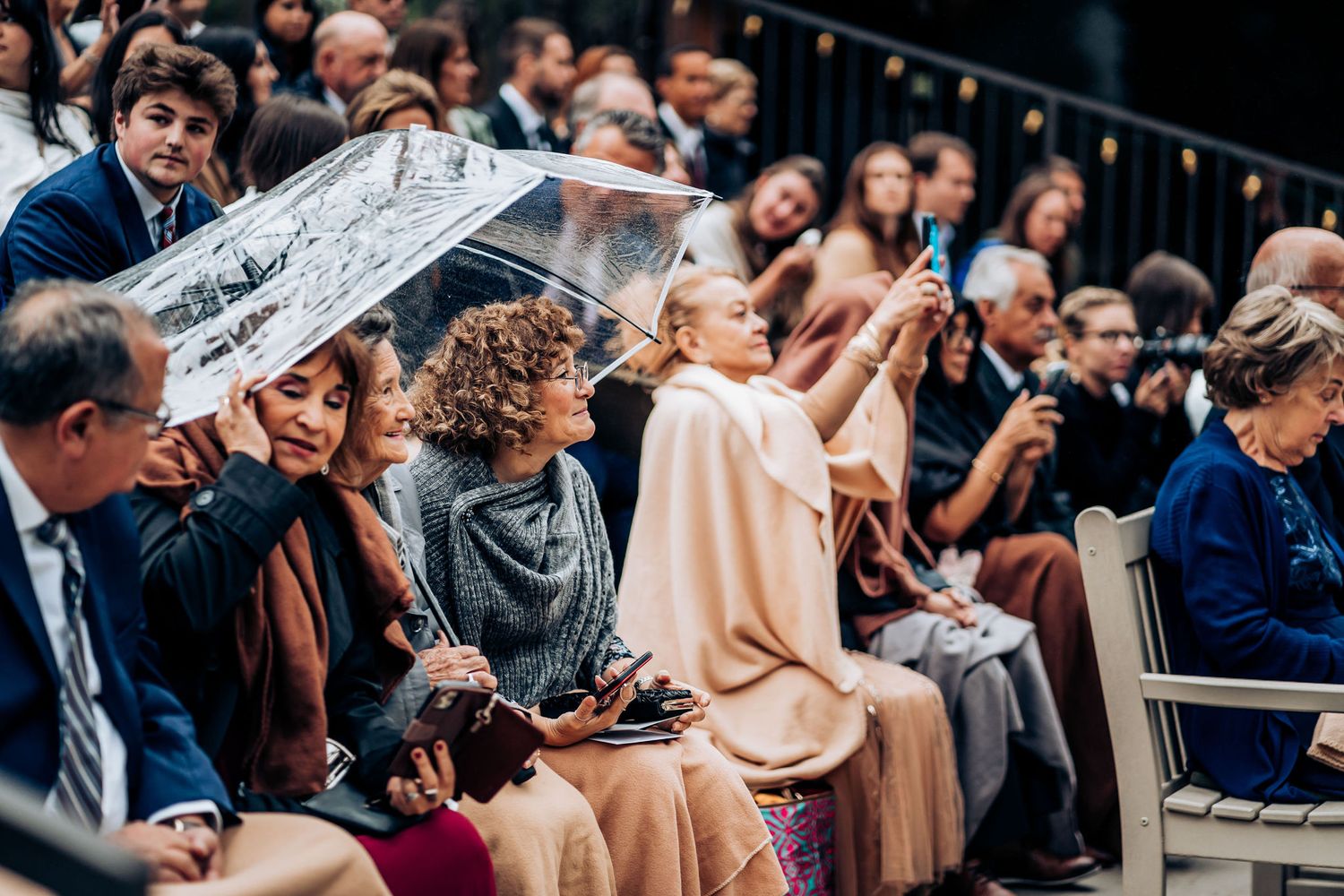 Wedding guests sheltering under clear umbrellas while watching ceremony in audience seating.