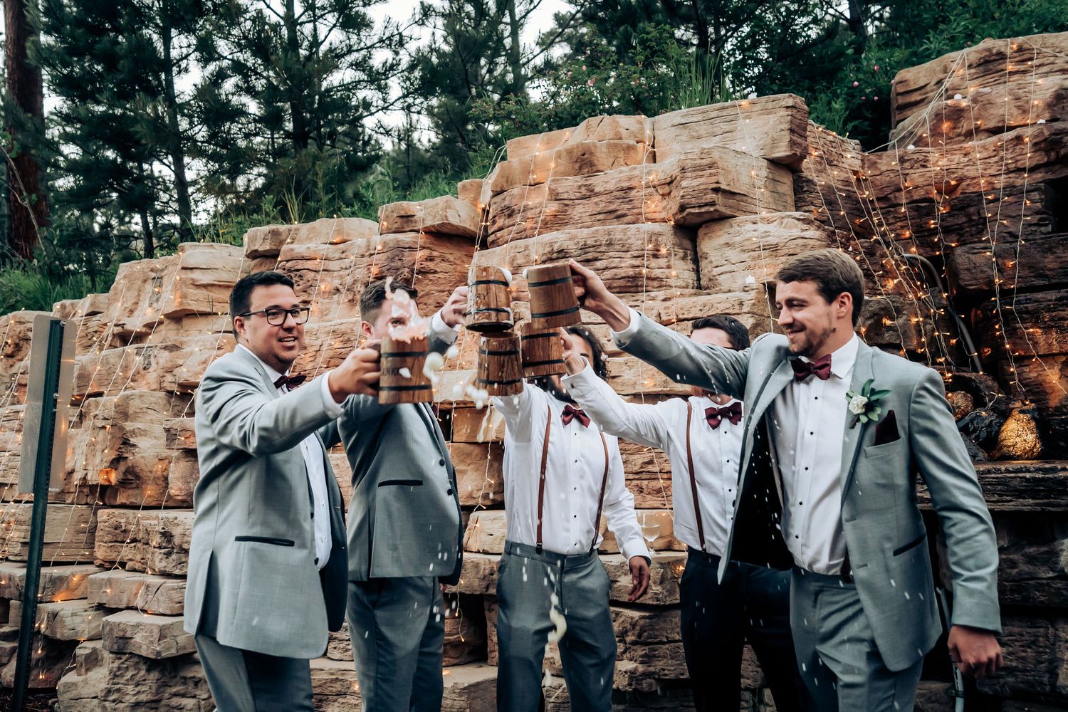 Groomsmen in grey suits pose playfully together against a stone wall backdrop.