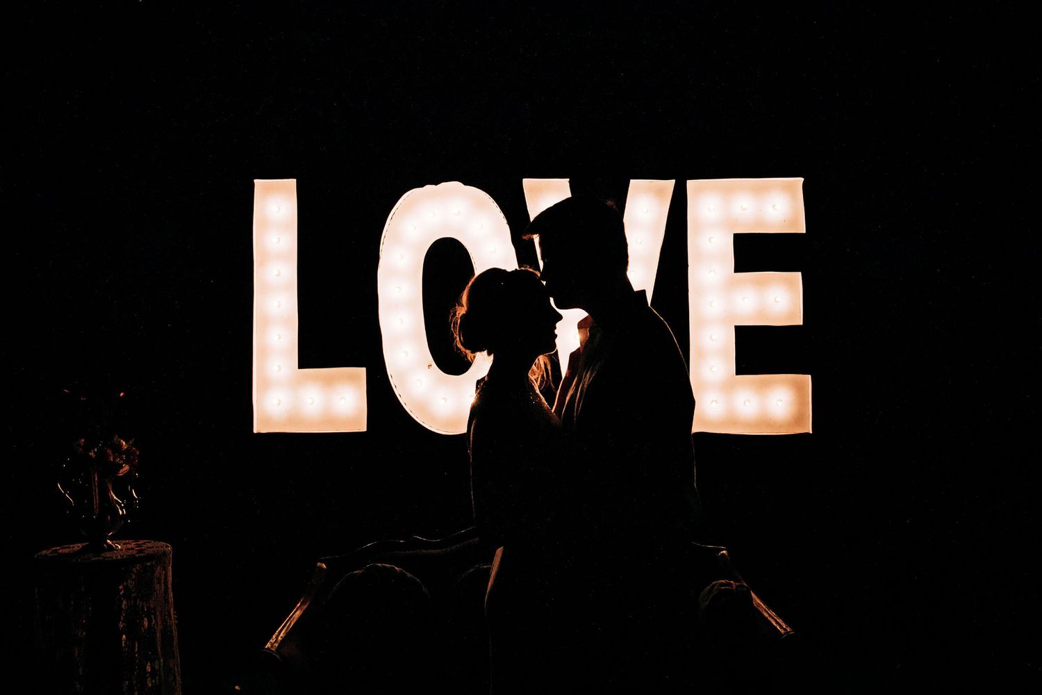 Romantic silhouette of couple against glowing LOVE sign at wedding reception.