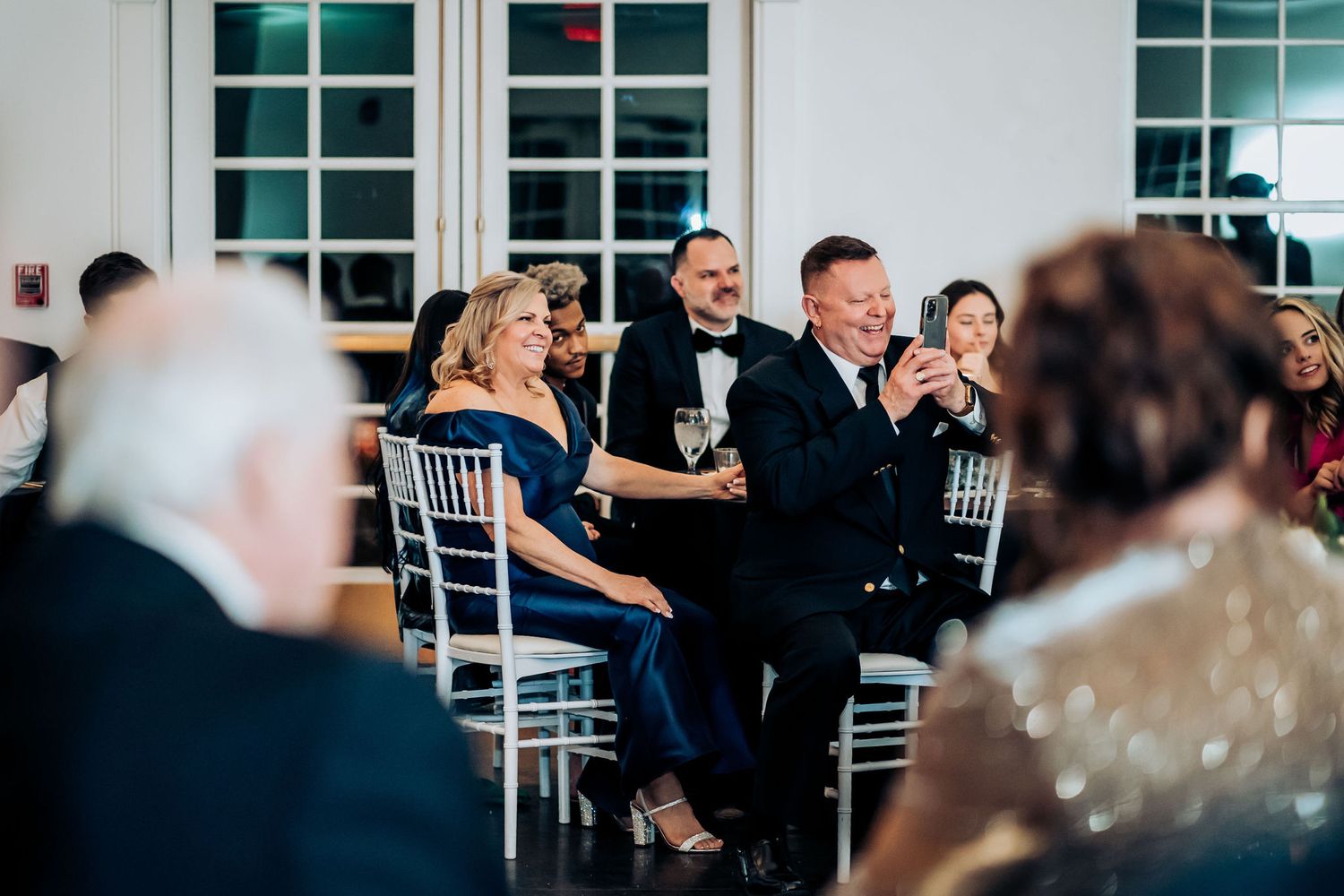 Wedding guests react during speeches at an elegant indoor reception with white chairs and decor.