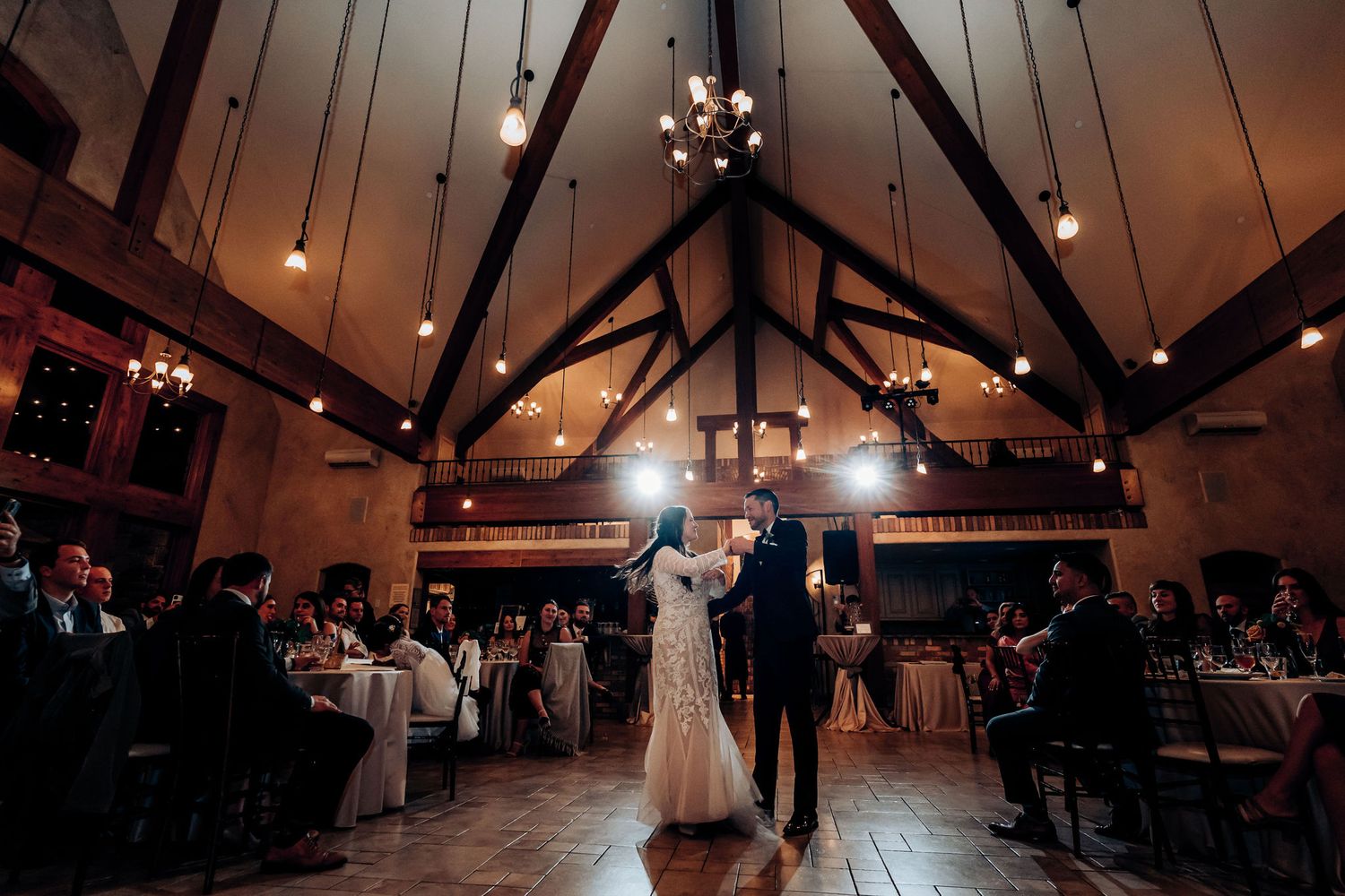 Wedding couple dances beneath dramatic vaulted ceilings with hanging lights in a rustic reception venue.