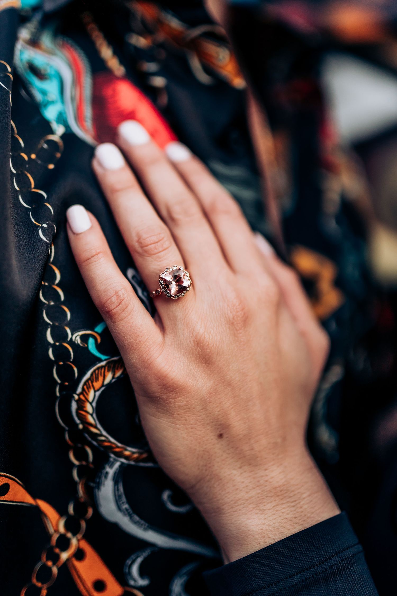 A close-up of a hand wearing an engagement ring against a colorful patterned background.