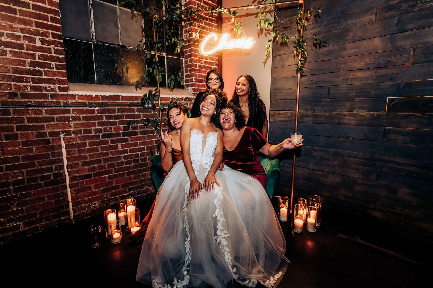 Bridal party gathers around illuminated signage with candles and exposed brick walls.