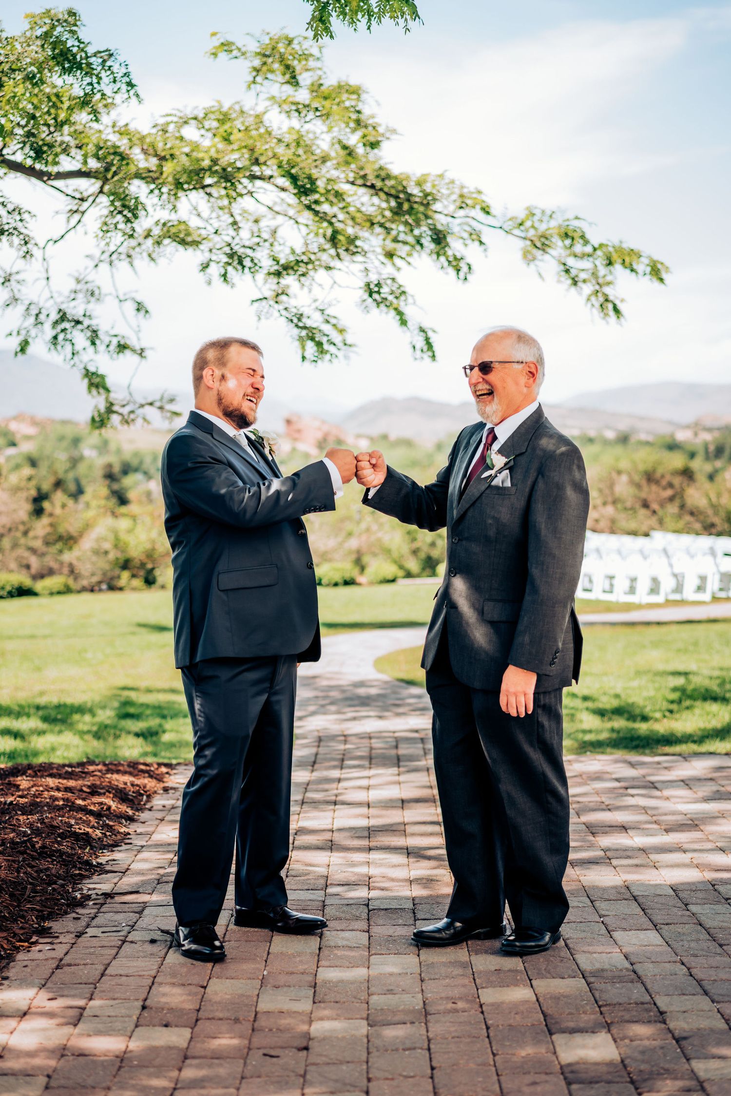 Two men in suits share a celebratory handshake on brick patio under tree branches.