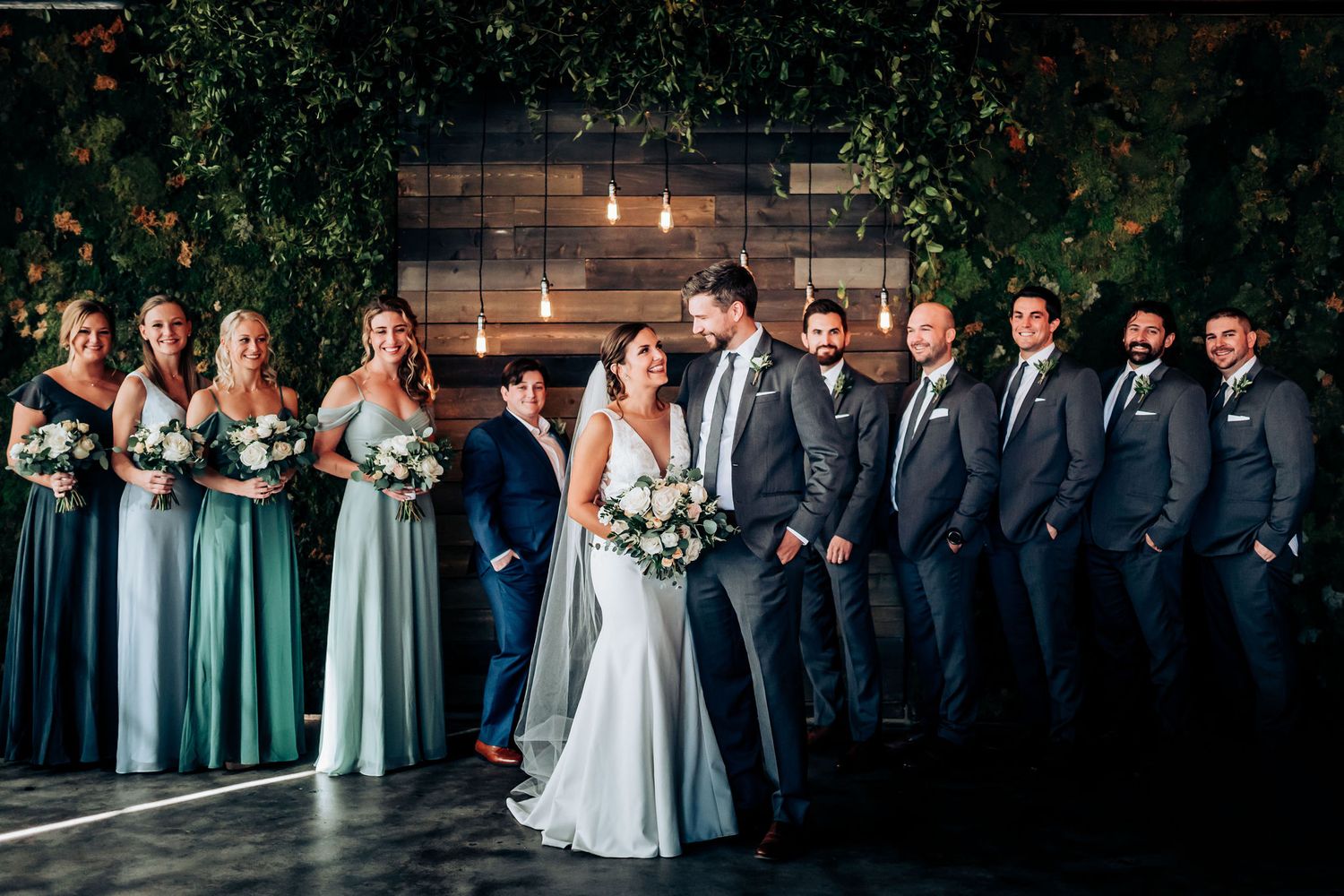 Wedding party poses for formal photo with bridesmaids in sage green dresses against rustic backdrop.