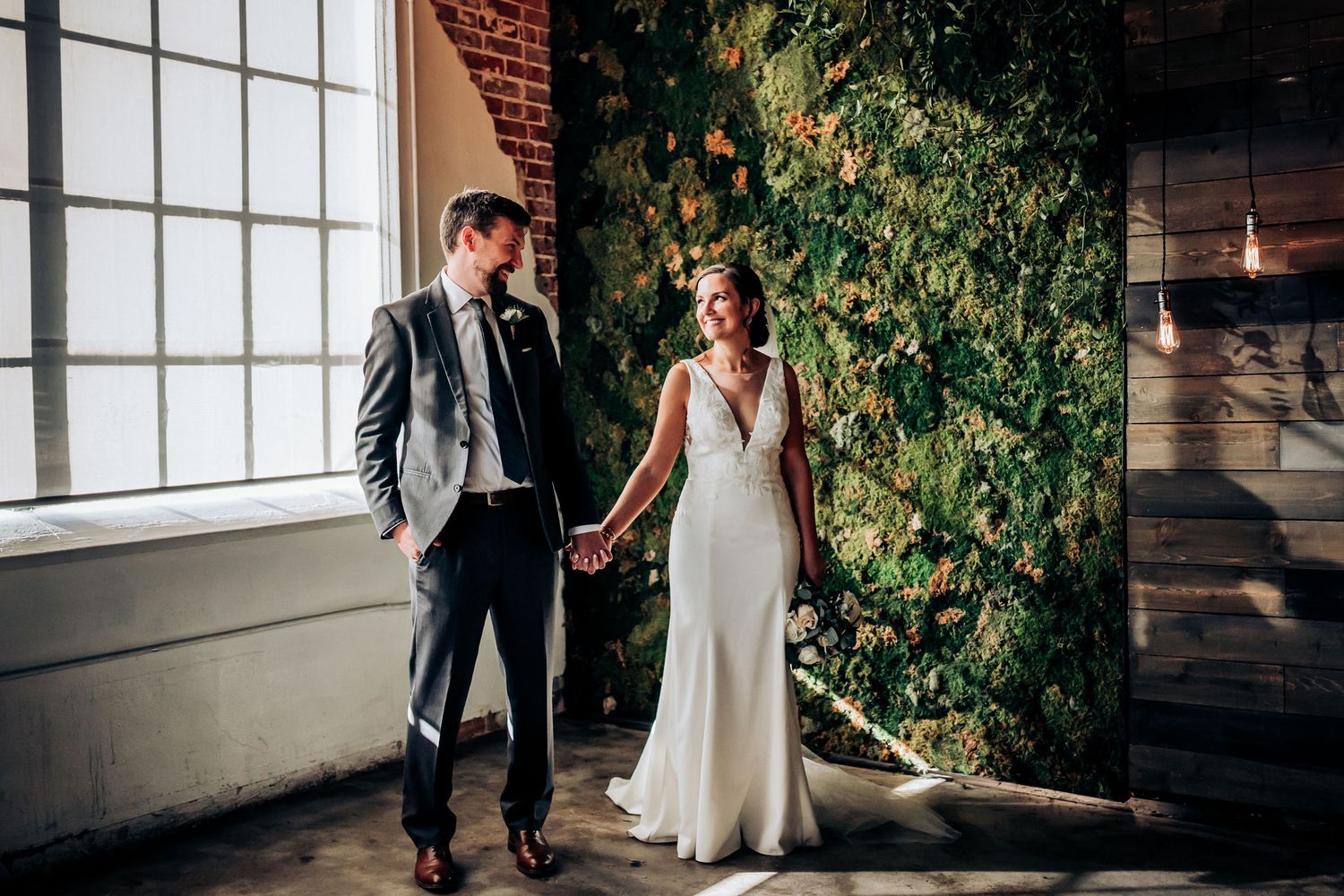 Couple in wedding attire standing by large windows and ivy-covered walls.