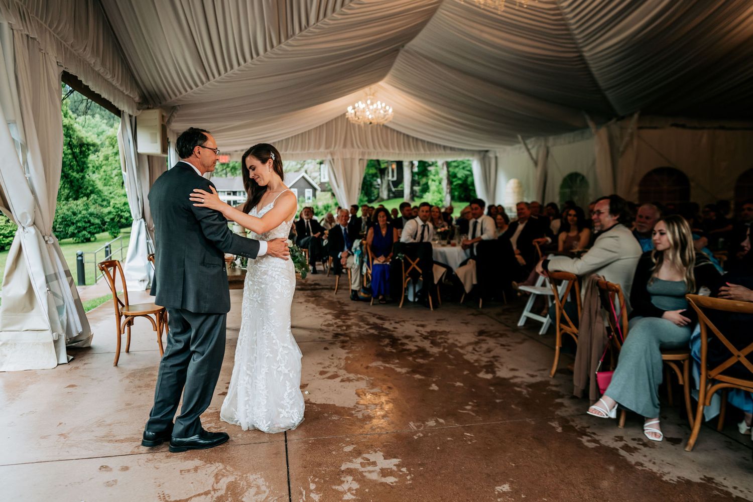 A couple shares their first dance at a wedding reception under an elegantly draped tent with guests watching.