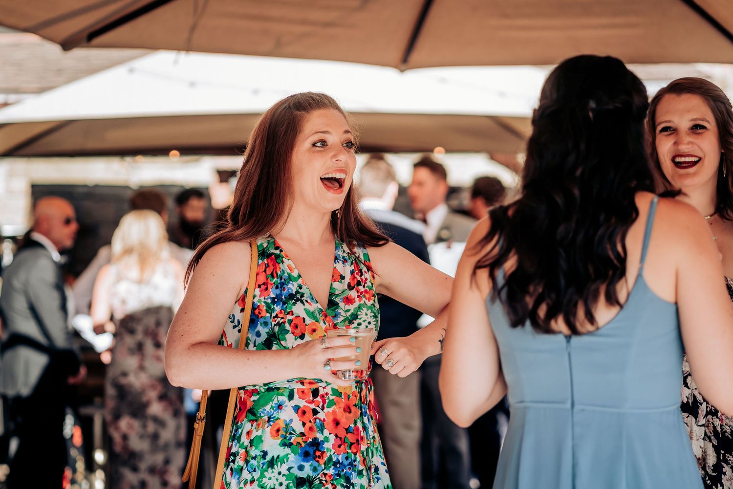 Two guests laugh and dance together under a brown umbrella during an outdoor wedding reception.