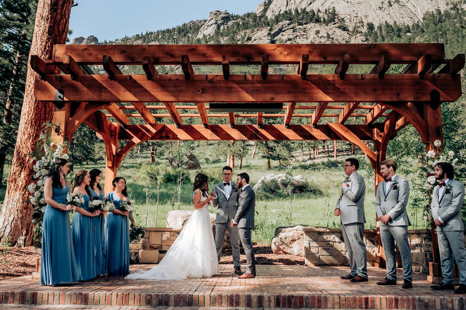 Outdoor mountain wedding ceremony under rustic wooden pergola with bridesmaids in blue dresses and groomsmen in grey suits.