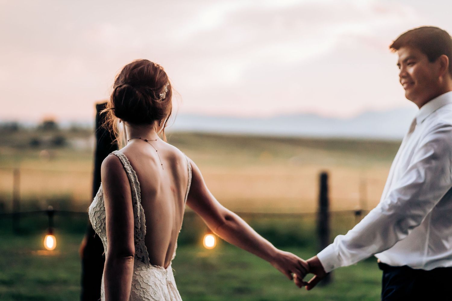 A couple dances intimately at sunset with string lights and rolling hills in the background.