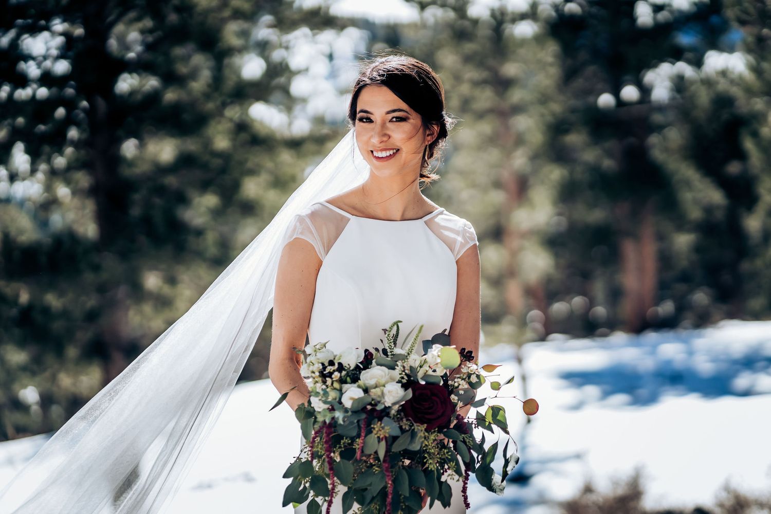 Elegant bride in white gown and flowing veil holds winter bouquet against snowy mountain backdrop.
