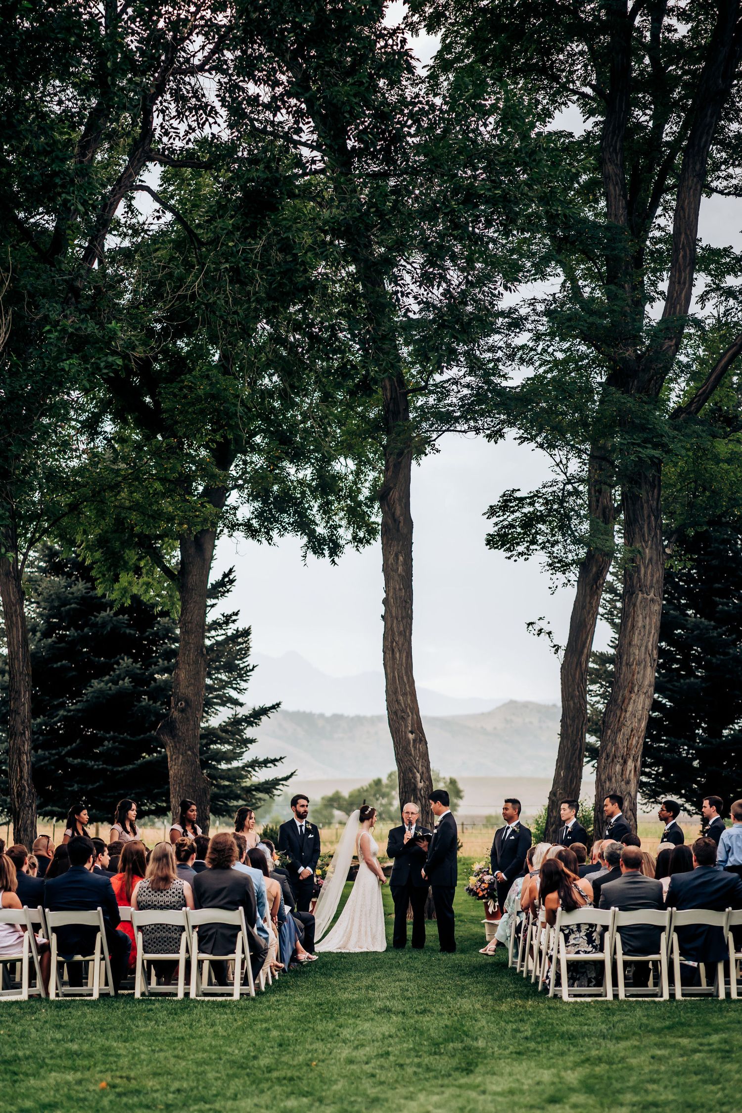 Outdoor wedding ceremony setup with white chairs framed by towering trees and mountain views in the background.