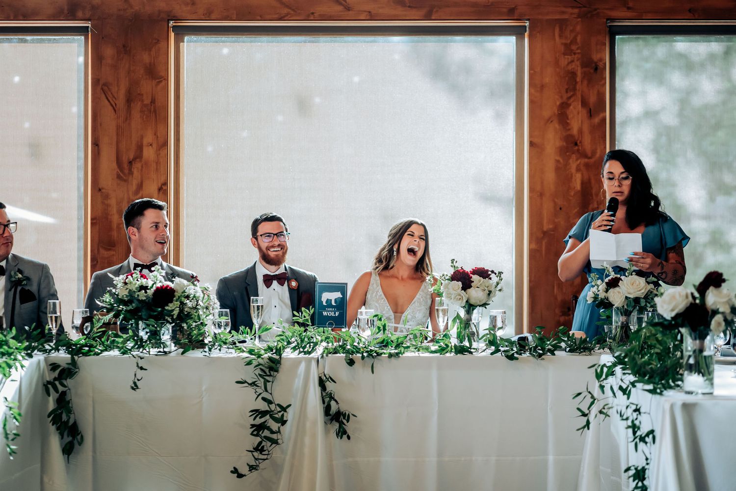 Wedding party sits at decorated head table with cascading greenery and white tablecloths.