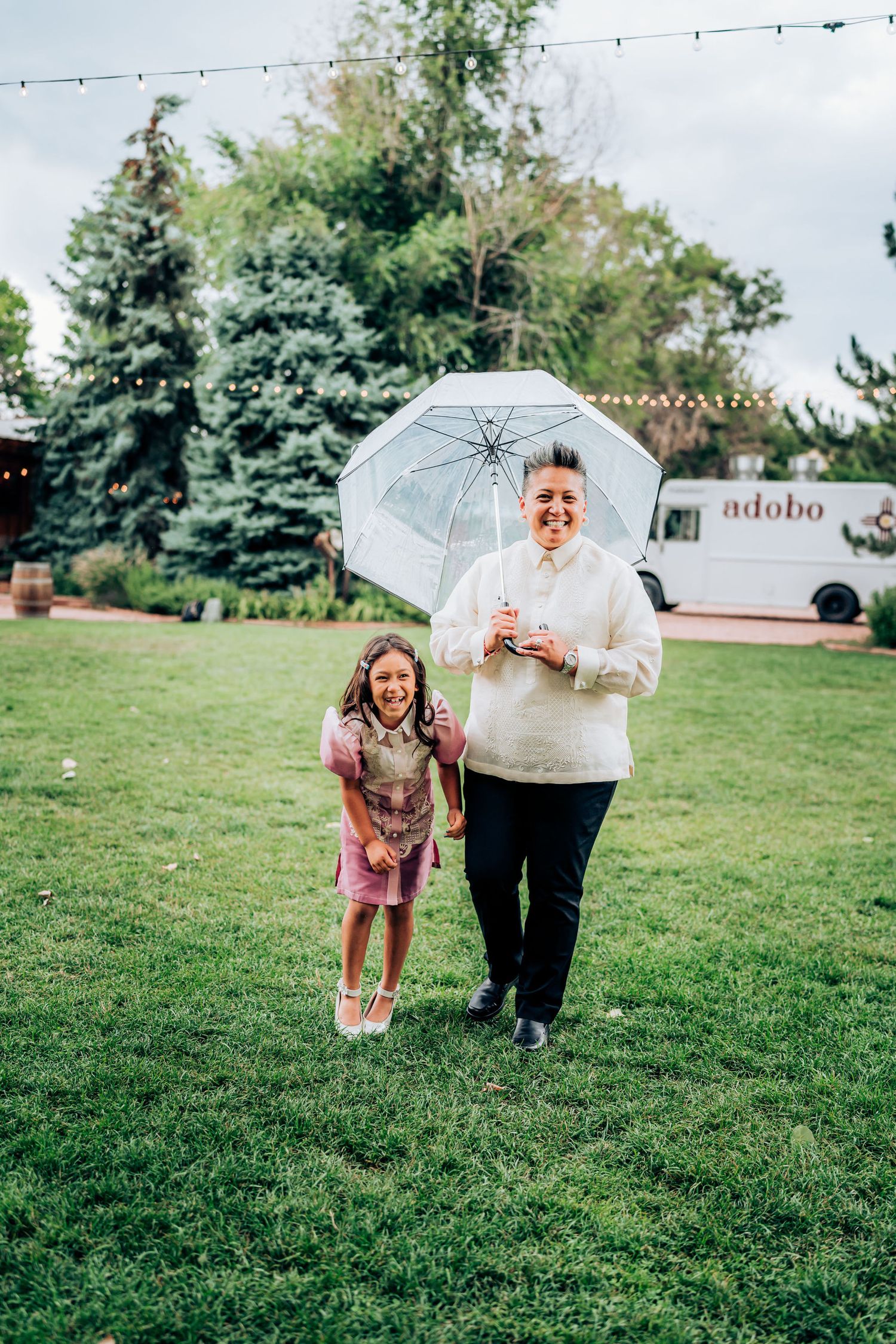 Joyful moment shared under a clear umbrella on a lawn with string lights and food truck visible.
