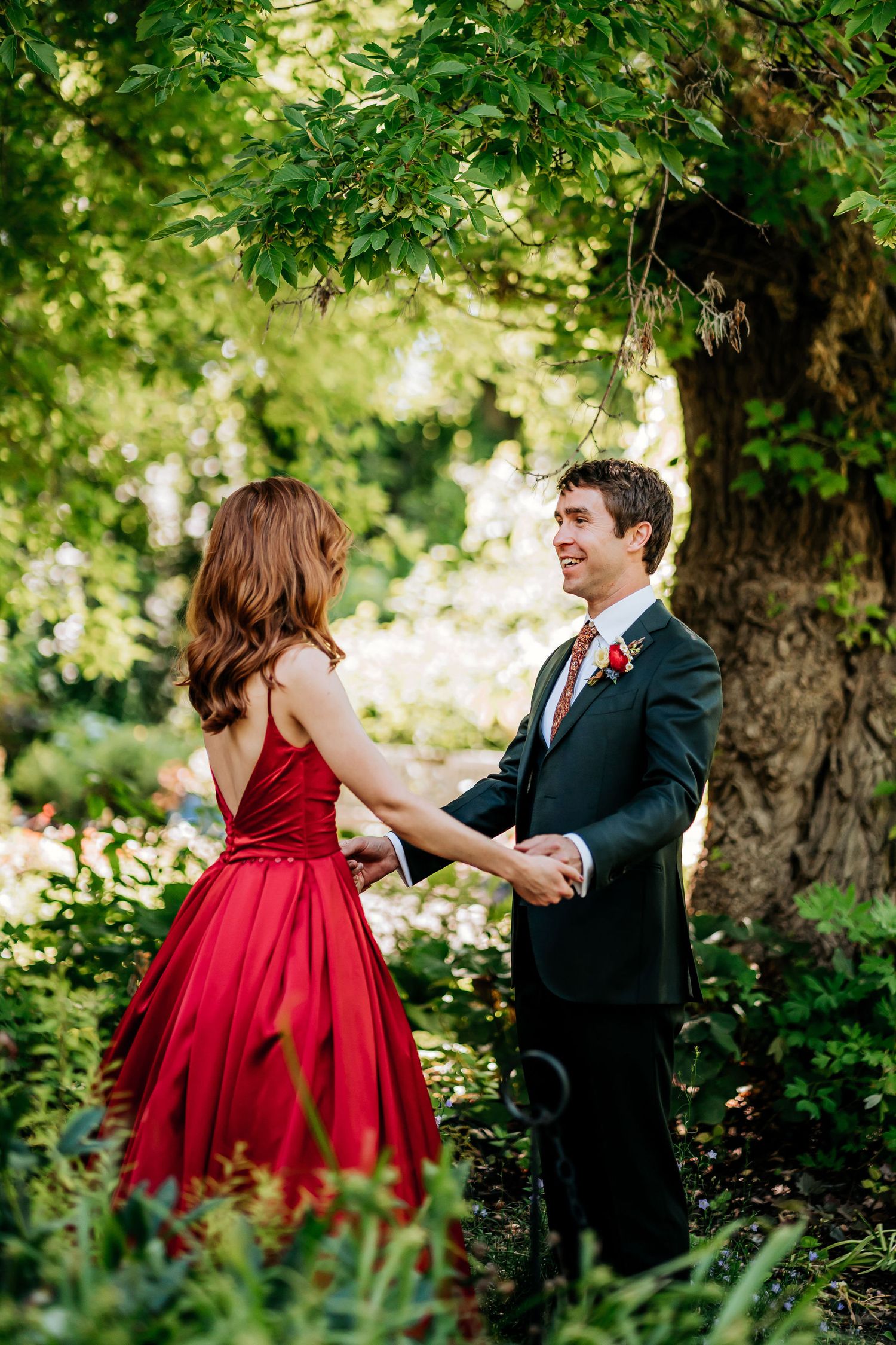 A couple shares a romantic moment in formal attire under a lush green tree in a garden setting.