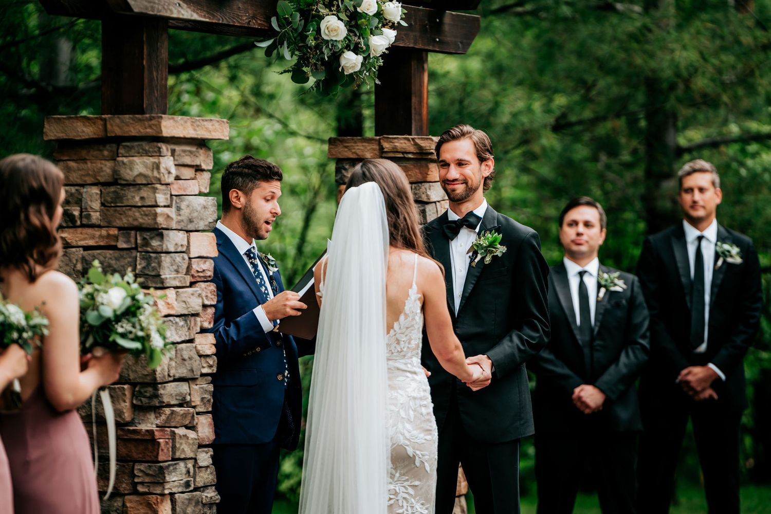 Outdoor wedding ceremony under stone archway with wedding party in formal attire.