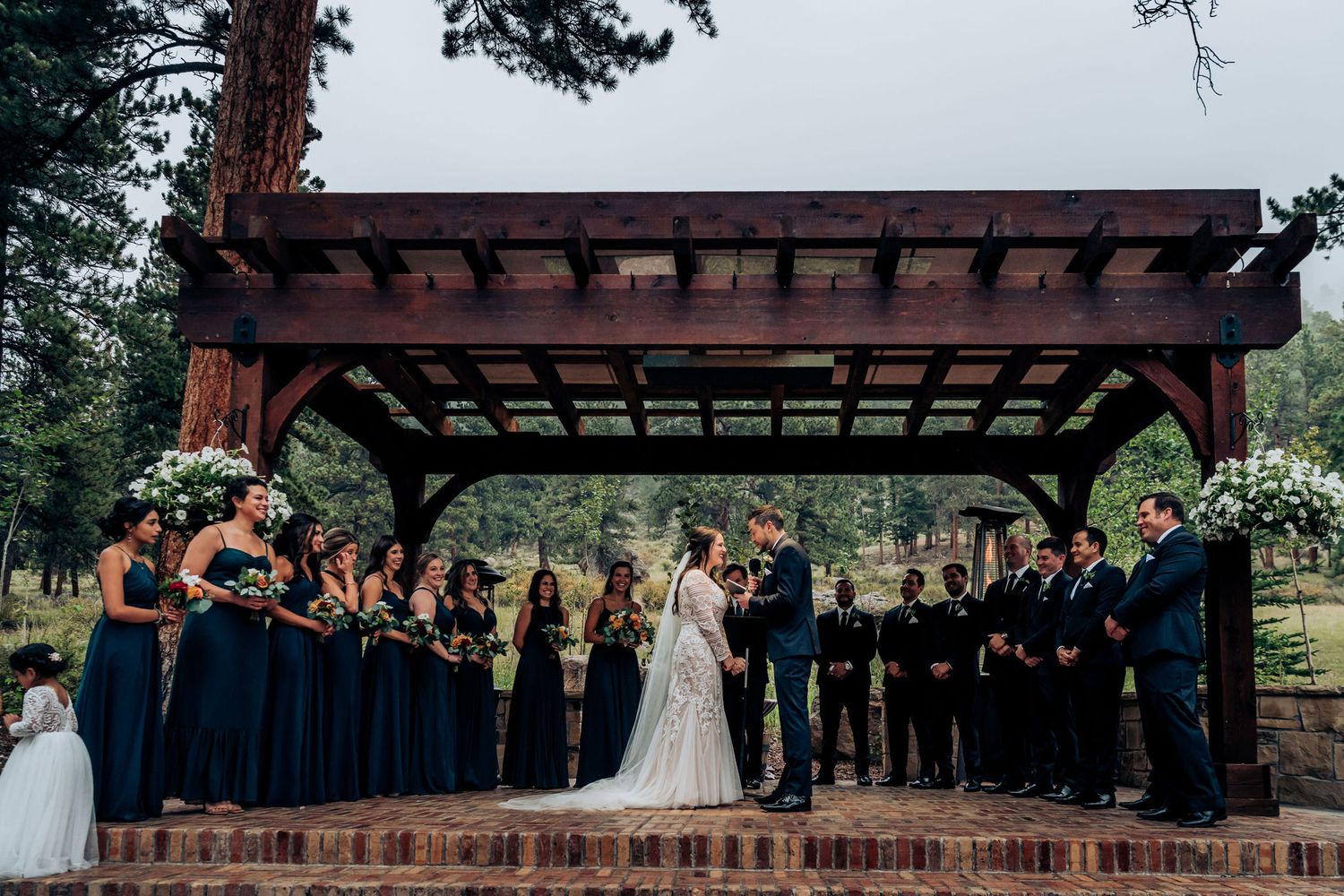 Wide shot of wedding ceremony with bridal party in navy and black lined up under wooden pergola.