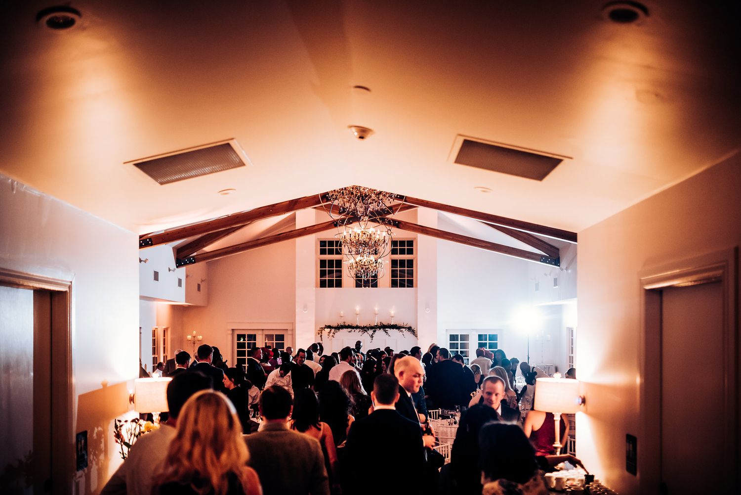 Wide shot of crowded wedding reception venue with warm ambient lighting and exposed beams.