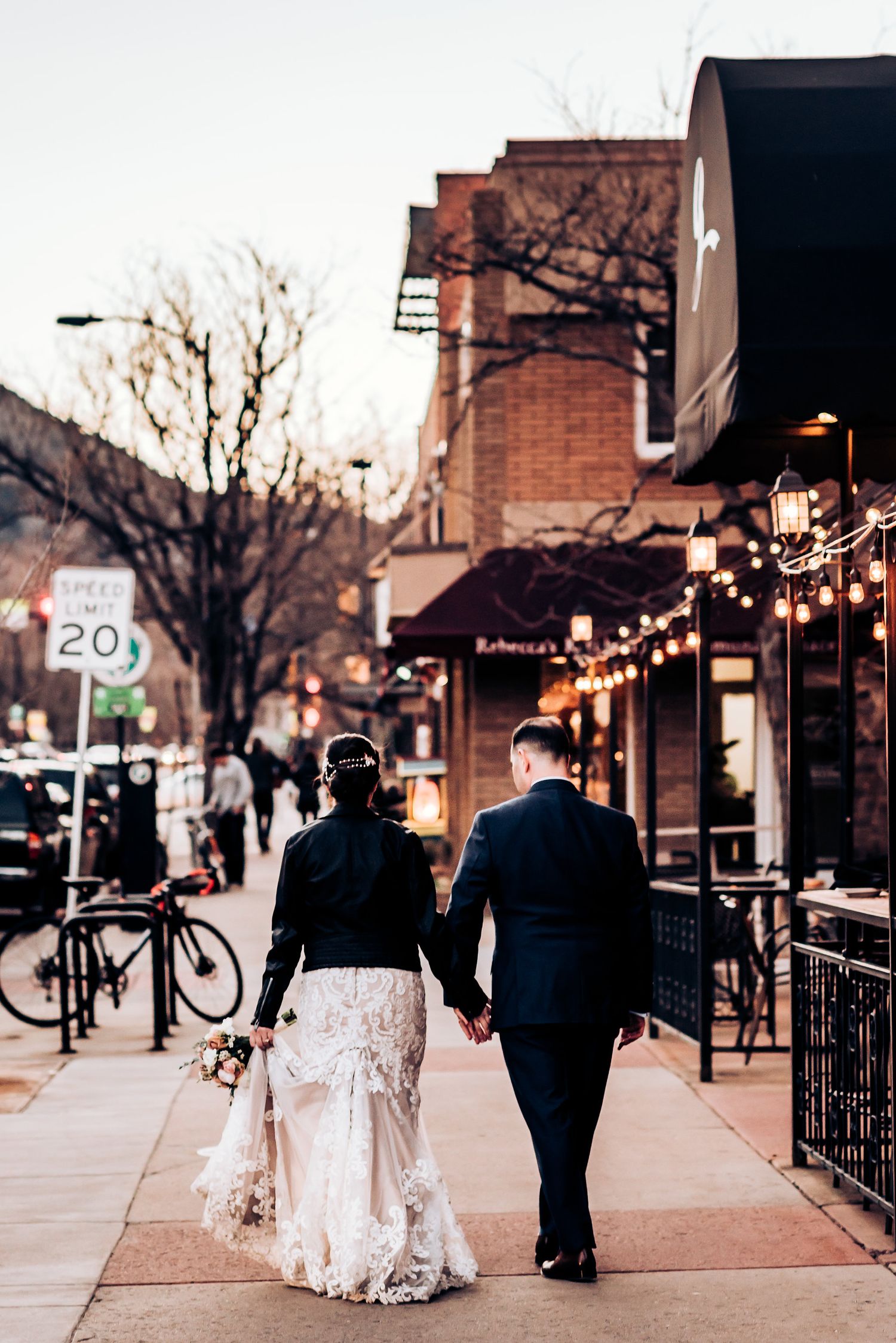 Newlyweds walking hand in hand down urban street with string lights and storefronts.