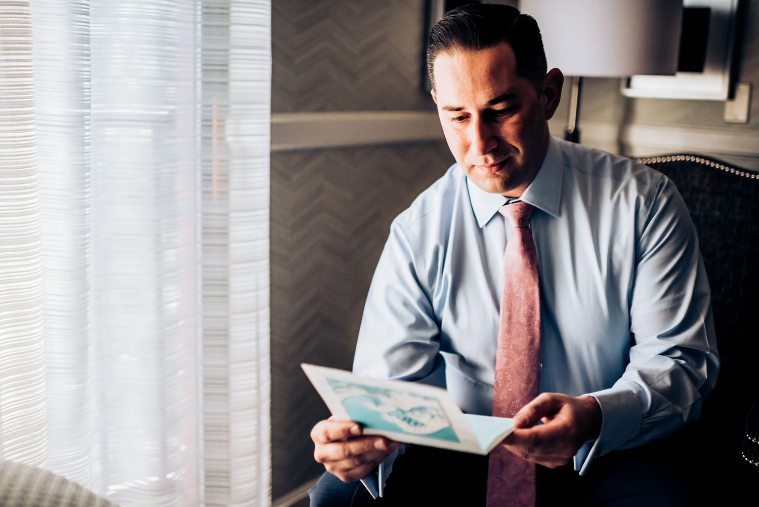 A businessman in a light shirt and pink tie reviews documents while seated by a window.