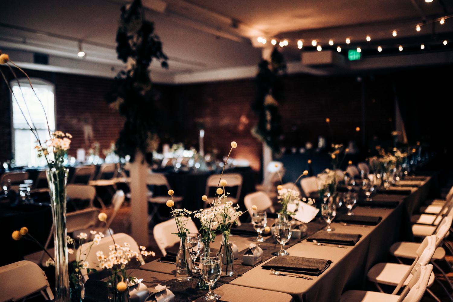 Long reception table decorated with elegant floral centerpieces and string lights in a rustic indoor venue.