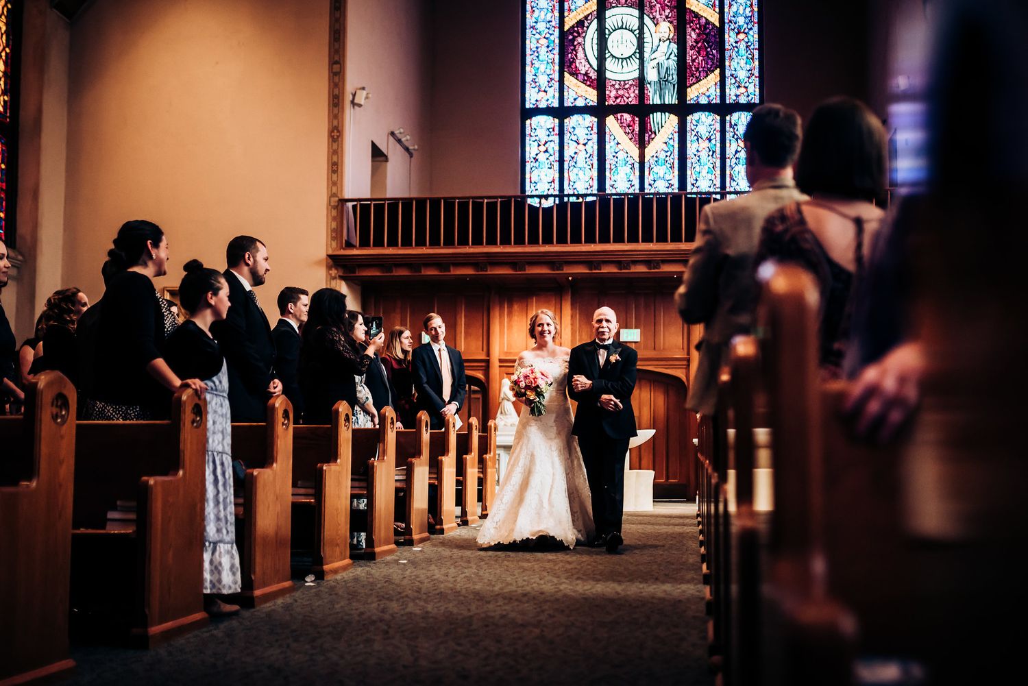 Church wedding ceremony with stained glass windows and guests seated in wooden pews.