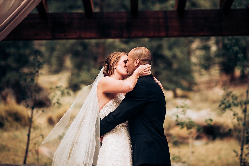 Romantic wedding moment captured under rustic wooden pergola with soft natural lighting.