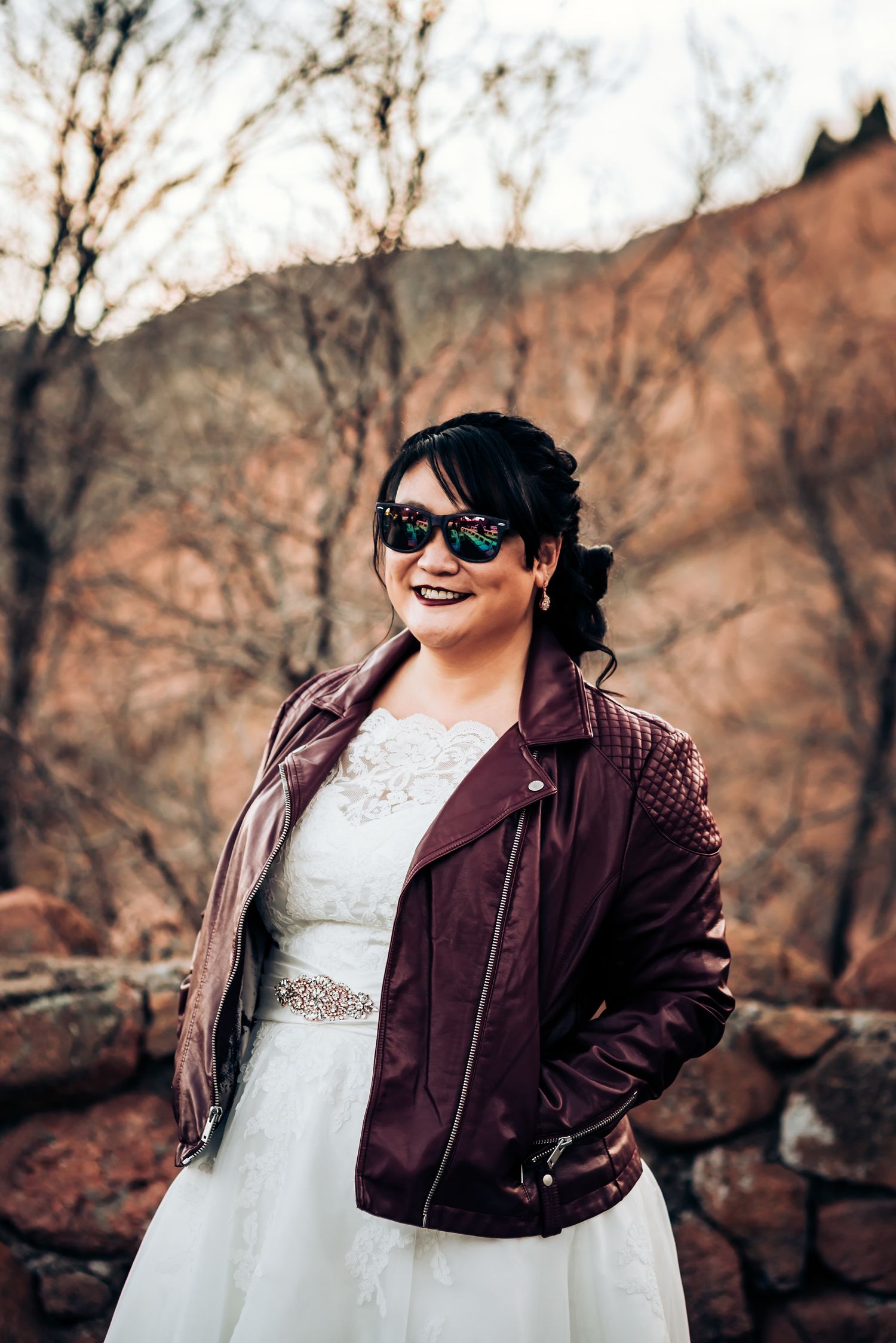 Bride in white dress and burgundy leather jacket poses confidently against desert backdrop.