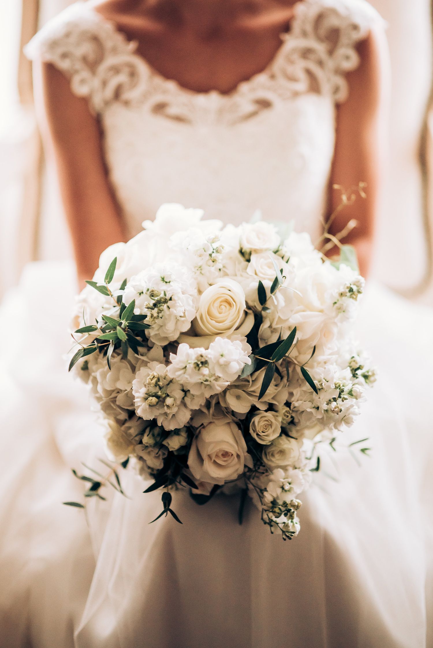 A stunning white wedding bouquet with roses, delicate lace details, and greenery held against a wedding dress.