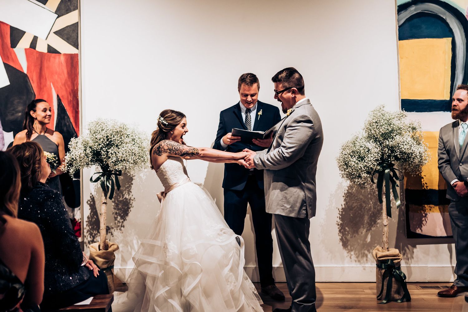 Wedding ceremony vows being exchanged in front of abstract wall art and floral arrangements.