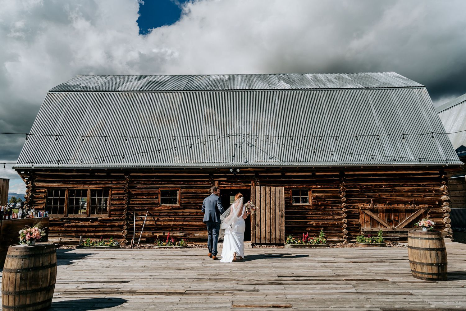 Rustic wooden barn with metal roof serves as backdrop for wedding photo.