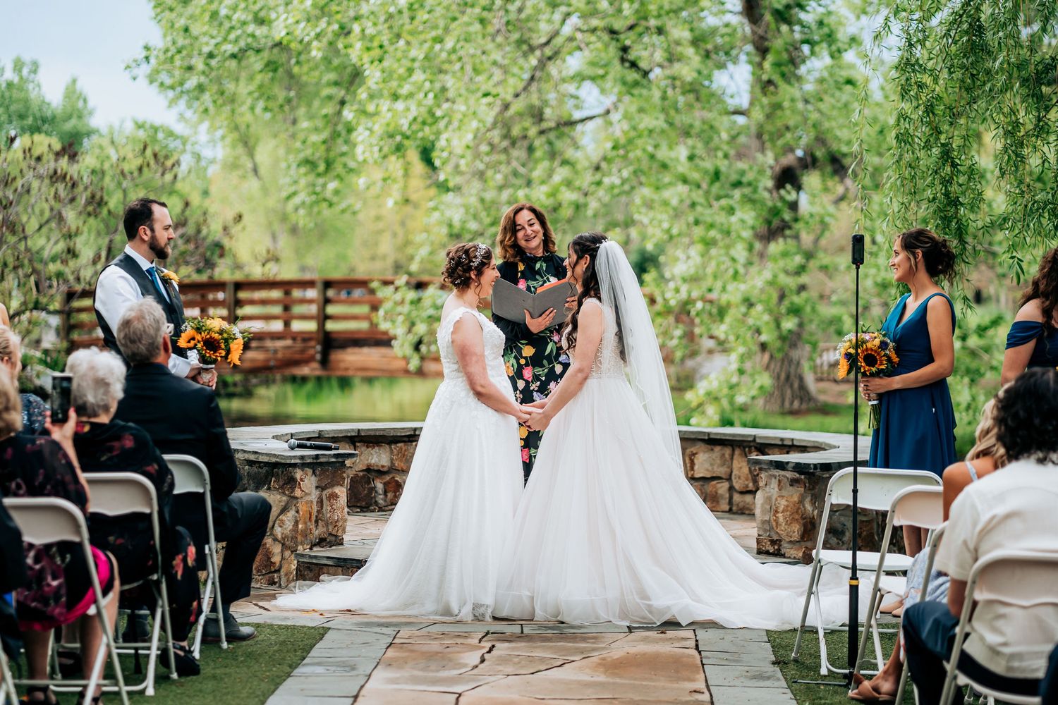 Wedding ceremony on a stone patio surrounded by lush green trees and white folding chairs.