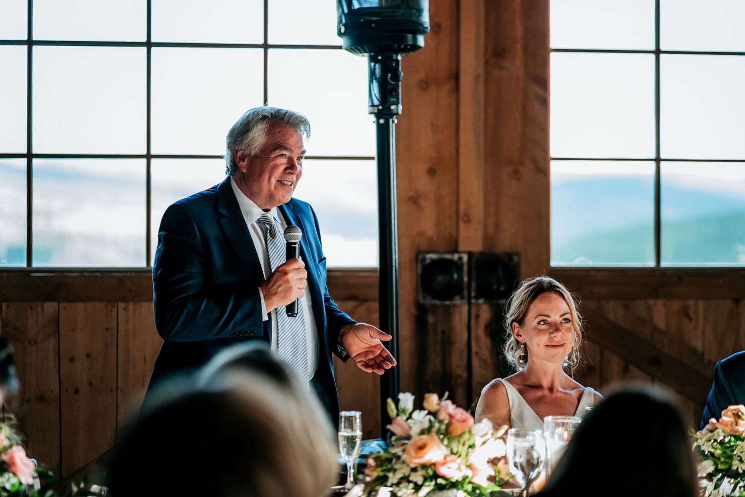 A speaker delivers a heartfelt toast at a formal dinner event in a rustic venue with large windows.