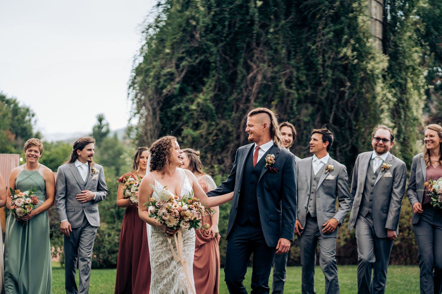 Wedding party laughs together during formal photos outdoors near vine-covered building.