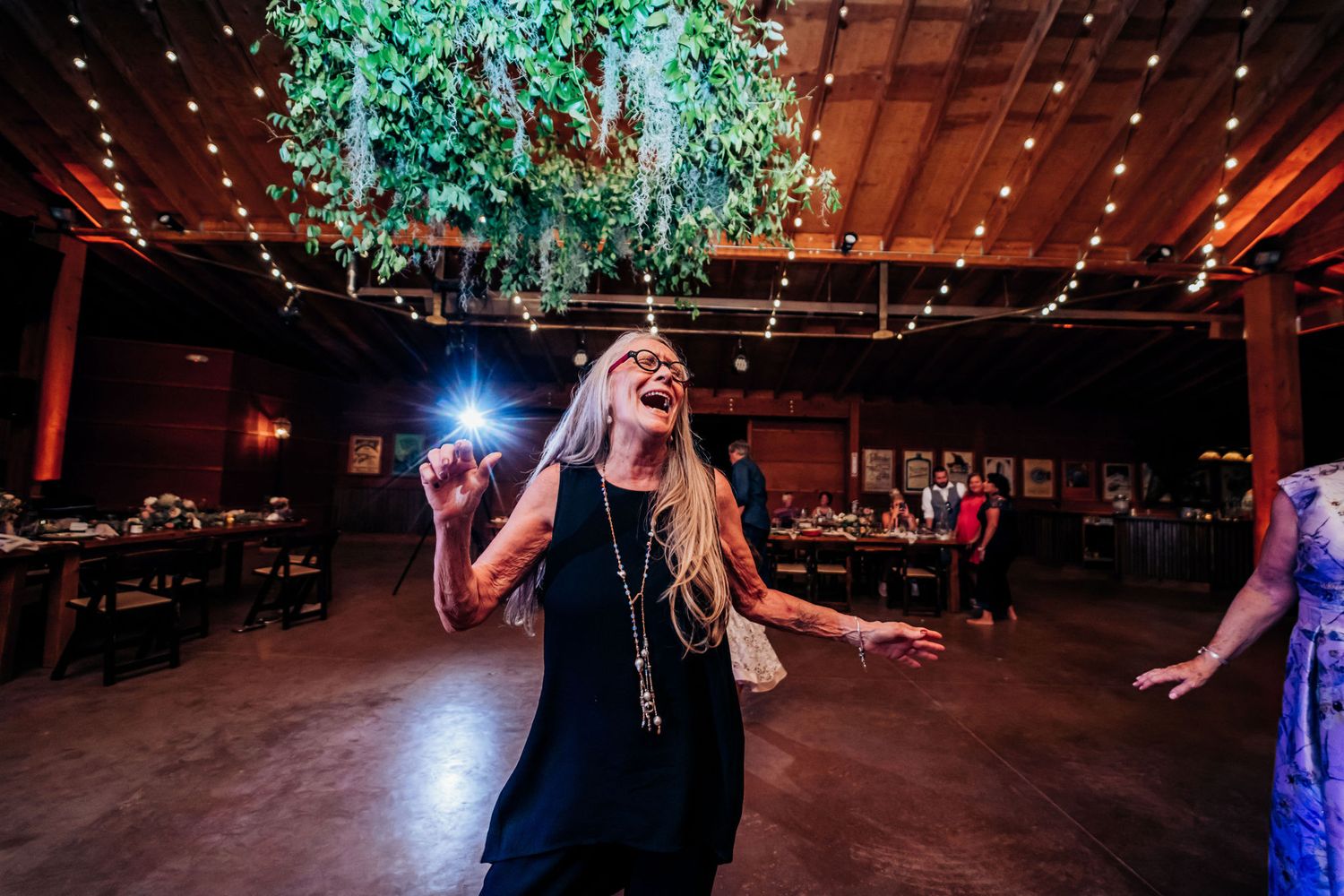 Woman dances joyfully under a hanging greenery installation at an indoor wedding reception.