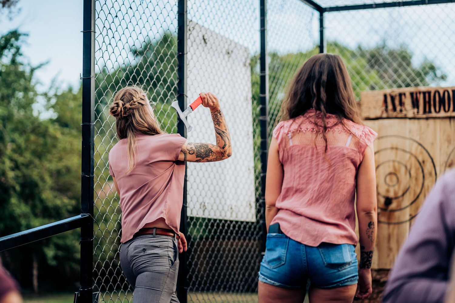 Two people in pink shirts participate in an axe throwing activity at an outdoor venue.